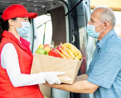 Delivery Man With Protection Mask Delivering Food To The Senior Man During Covid19 Pandemic Or Lockdown.